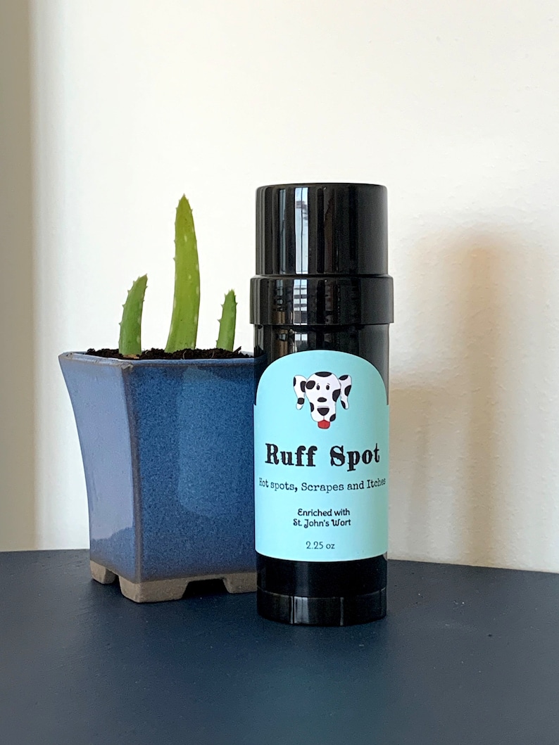 May include: A black cylindrical container of "Ruff Spot" for pets, with a light blue label featuring a Dalmatian illustration. The label reads "Hot spots, Scrapes and Itches" and "Enriched with St. John's Wort", 2.25 oz. A blue square pot with aloe vera is in the background.