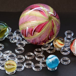 May include: A collection of colorful glass marbles displayed on clear plastic rings. The marbles are various sizes and feature different patterns, including swirls, stripes, and solid colors. The largest marble is a pink and green swirl.
