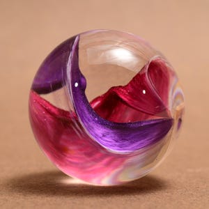 Peut inclure: Une sphère en verre transparent avec des motifs tourbillonnants intérieurs dans des tons de violet, de rose et de rouge. La sphère a une surface lisse et polie et un centre creux, créant un objet décoratif unique.