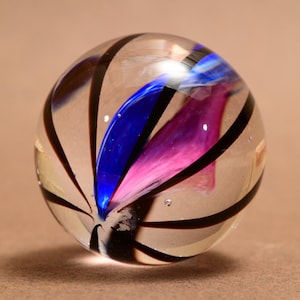 Könnte beinhalten: Eine transparente Glaskugel mit schwarzen, gebogenen Streifen, die von der Mitte ausgehen. Im Inneren der Murmel befinden sich blaue und rosa Wirbel. Die Murmel ist eine Kugel.