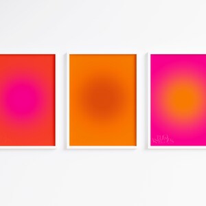 May include: Three framed abstract art prints with a gradient design. The prints feature a blend of vibrant colors: red and pink, orange, and pink and orange. The artwork is displayed on a white wall.
