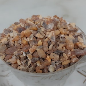 Puede incluir: Una taza de porcelana blanca llena de pequeños trozos irregulares de resina de color ámbar. La resina es una mezcla de tonos marrón claro y oscuro.