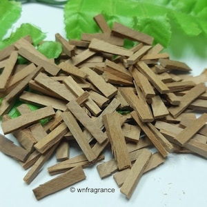 May include: A pile of small, rectangular, light brown wood pieces, possibly incense or fragrance sticks, are scattered on a white surface. Green leaves are in the background. The image is well-lit, highlighting the natural wood grain.