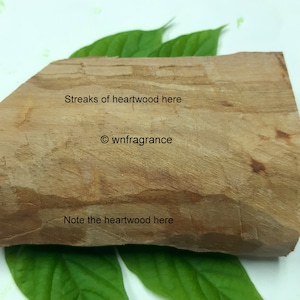 May include: A light brown heartwood block, showing streaks and grain, is displayed on green leaves. The text "Streaks of heartwood here", "© wnfragrance", and "Note the heartwood here" are visible on the wood.
