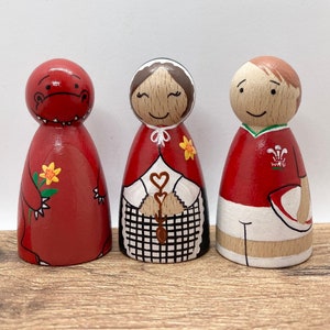 May include: Three wooden peg dolls, painted in red, white, and black. The doll on the left is a red dragon, the middle doll is a woman wearing a red dress and a black and white checkered apron, and the doll on the right is a man wearing a red rugby jersey with the Welsh flag on it.