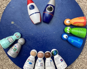 May include: A wooden space-themed toy set featuring a blue circular base with painted stars. The set includes two rocket ships, several astronaut figures, and alien figures. The rockets are red, white, and blue. The astronauts are in white space suits.