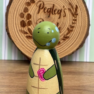 May include: A hand-painted wooden turtle peg doll with a green head and a beige body with a painted shell design. The turtle holds a pink heart-shaped object. The background includes a wooden slice with the text "Pegleg's".