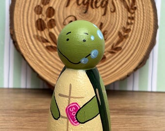 May include: A hand-painted wooden turtle peg doll with a green head and a beige body with a painted shell design. The turtle holds a pink heart-shaped object. The background includes a wooden slice with the text "Pegleg's".