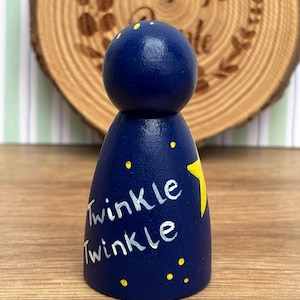 May include: A dark blue wooden peg doll with a round head and a conical body. The body is decorated with a yellow star and the words "Twinkle Twinkle" in white. Small yellow dots are scattered around the body.