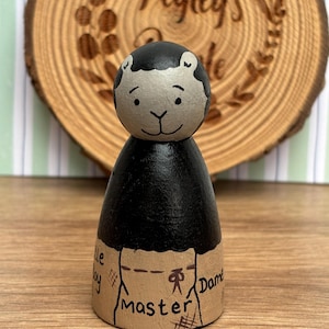 May include: A hand-painted wooden peg doll with a black body and a light gray head. The head has black hair and a smiling face. The doll's base is tan with the word "Master" and other text. The background is a wooden slice with the text "Pegleg's Made".