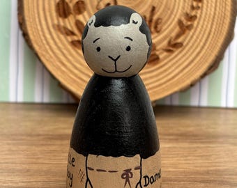 May include: A hand-painted wooden peg doll with a black body and a light gray head. The head has black hair and a smiling face. The doll's base is tan with the word "Master" and other text. The background is a wooden slice with the text "Pegleg's Made".