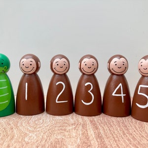 May include: A set of six wooden peg dolls. One is a green alligator, and the other five are brown monkeys with white numbers 1-5. The dolls have smiling faces and are painted with non-toxic paint.