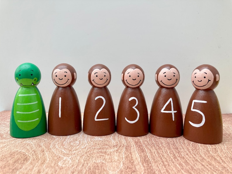 May include: A set of six wooden peg dolls. One is a green alligator, and the other five are brown monkeys with white numbers 1-5. The dolls have smiling faces and are painted with non-toxic paint.