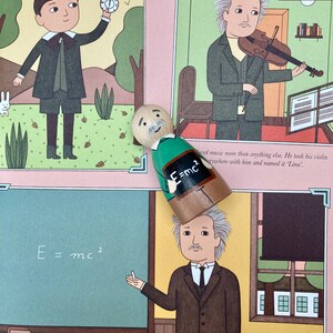 May include: A wooden peg doll with a painted Einstein design, standing over a book illustration. The illustration depicts Einstein at different ages, including one with the equation E=mc&sup2;. The background is a light pink color.