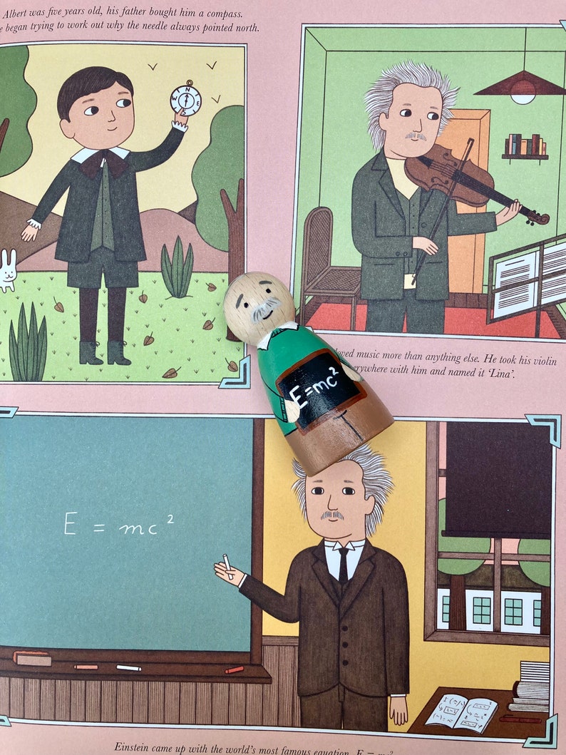 May include: A wooden peg doll with a painted Einstein design, standing over a book illustration. The illustration depicts Einstein at different ages, including one with the equation E=mc&sup2;. The background is a light pink color.