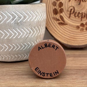 May include: A small, round, terracotta-colored object with the name "ALBERT EINSTEIN" in black lettering. The object is resting on a wooden surface, with a decorative pot and a wooden slice in the background.