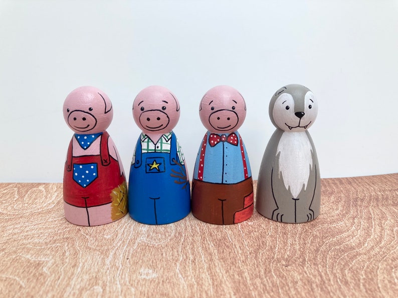 May include: Four wooden peg dolls, painted to represent characters from the story of the Three Little Pigs. The dolls are standing on a wooden surface. The dolls are painted in various colors, including pink, red, blue, brown, and gray. The dolls have painted facial features and clothing.