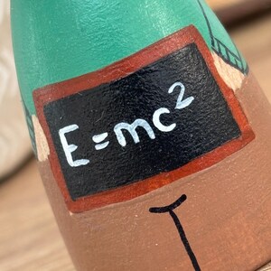 May include: A handcrafted wooden figurine of a person, painted with a green shirt and brown pants. A small chalkboard displays the equation E=mc&sup2;. The figurine has a white collar and a black tie.