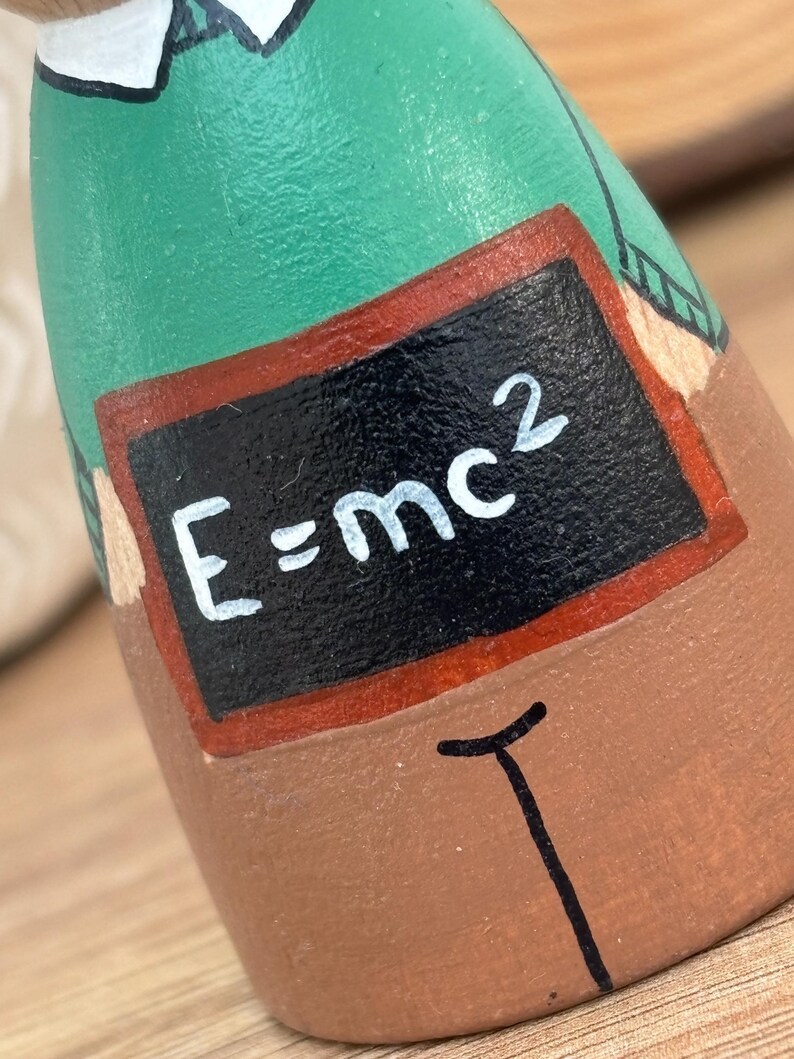 May include: A handcrafted wooden figurine of a person, painted with a green shirt and brown pants. A small chalkboard displays the equation E=mc&sup2;. The figurine has a white collar and a black tie.