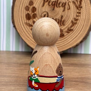 May include: A wooden peg doll with a rounded head and painted details. The doll features a cat and dog in a red boat on a blue base. The background includes a wooden slice with the text "Pegleg's People".