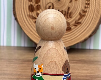 May include: A wooden peg doll with a rounded head and painted details. The doll features a cat and dog in a red boat on a blue base. The background includes a wooden slice with the text "Pegleg's People".
