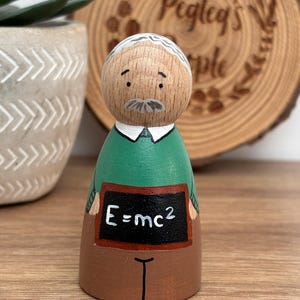 May include: A wooden peg doll depicting a scientist with a white mustache and hair. The doll wears a green shirt, brown pants, and holds a chalkboard with the equation E=mc&sup2;. A wooden slice with the text "Pegleg's People" is in the background.