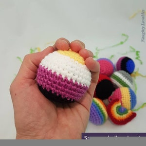May include: A crocheted ball with yellow, white, pink and black stripes. The ball is being held in a hand. There are other crocheted balls in the background, including a rainbow-coloured ball.