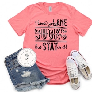 May include: A pink t-shirt with the text "I know your LANE SUCKS but STAY in it!" printed in black. The shirt is shown on a model wearing denim shorts and white trainers.