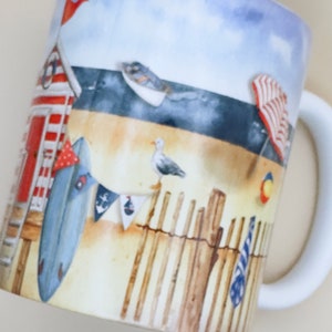 May include: A white ceramic mug with a beach scene design. The mug features a red and white striped beach hut, a blue surfboard, a seagull, and a wooden fence. The background is a blue sky with white clouds.