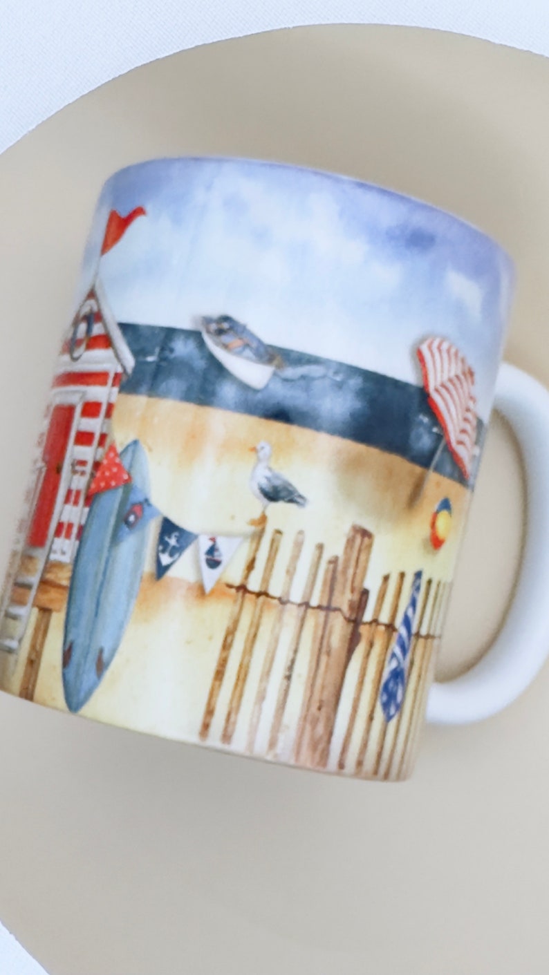 May include: A white ceramic mug with a beach scene design. The mug features a red and white striped beach hut, a blue surfboard, a seagull, and a wooden fence. The background is a blue sky with white clouds.