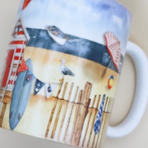 May include: White ceramic mug with a beach scene design. The mug features a red and white striped beach hut, a blue surfboard, a seagull, and a wooden fence. The background is a blue sky with white clouds.