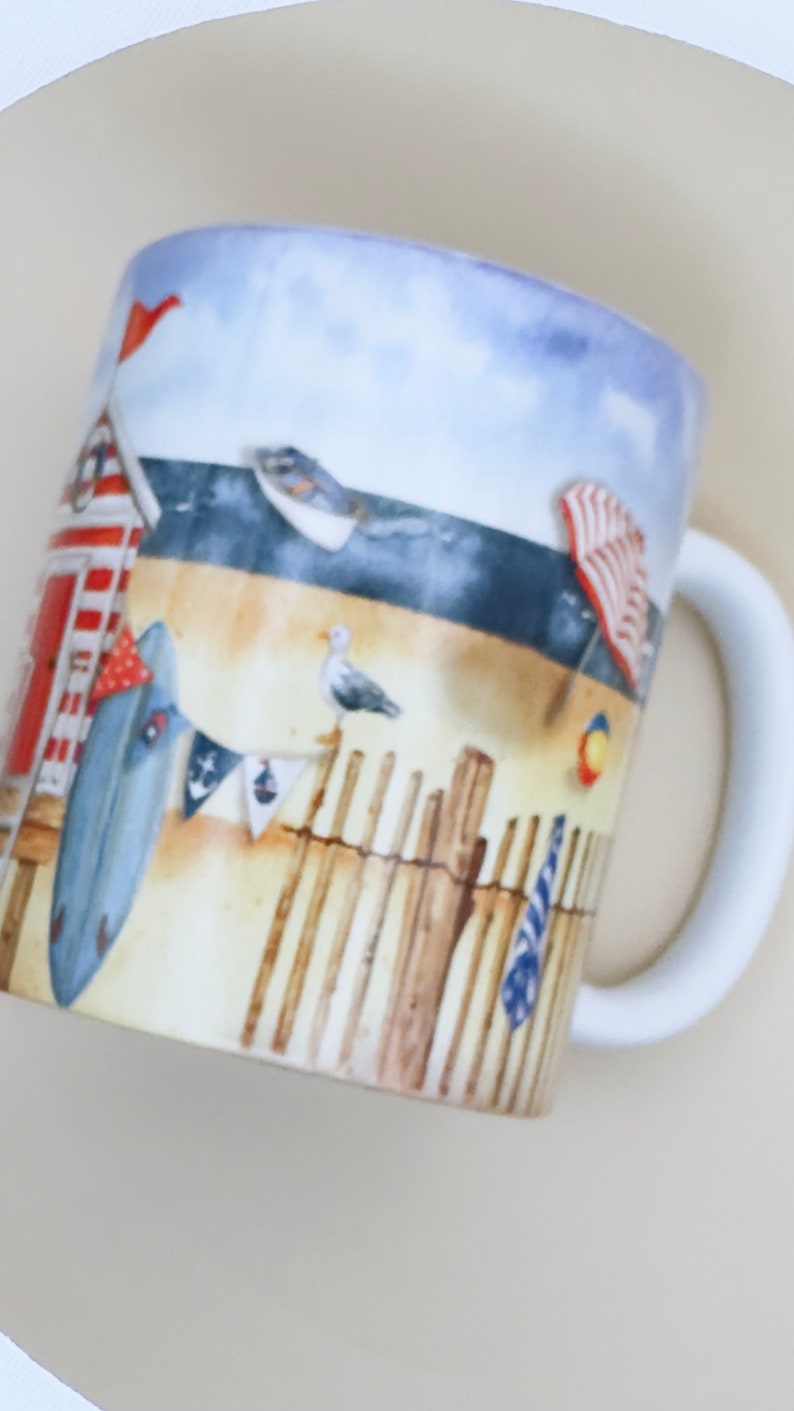 May include: White ceramic mug with a beach scene design. The mug features a red and white striped beach hut, a blue surfboard, a seagull, and a wooden fence. The background is a blue sky with white clouds.