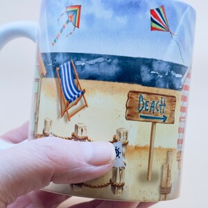 May include: A white ceramic mug with a beach scene design. The mug features a blue and white striped deckchair, a sign that says "Beach", and two colourful kites in the sky.