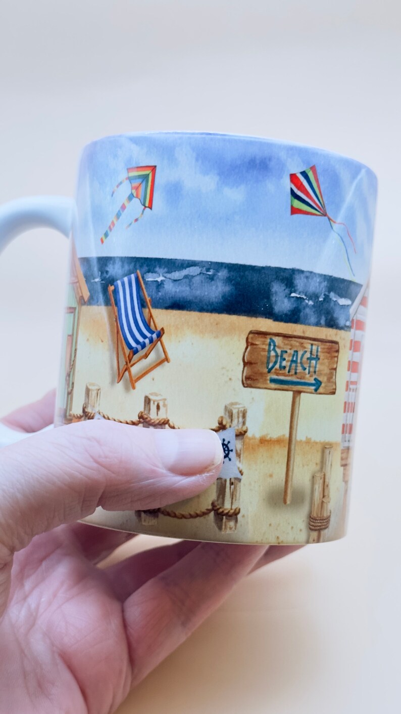May include: A white ceramic mug with a beach scene design. The mug features a blue and white striped deckchair, a sign that says "Beach", and two colourful kites in the sky.