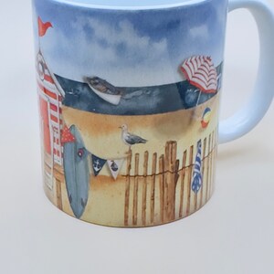 May include: A white ceramic mug with a beach scene design. The mug features a red and white striped beach hut, a blue surfboard, a red and white striped umbrella, a blue and white striped towel, and a seagull. The background is a blue sky with white clouds.