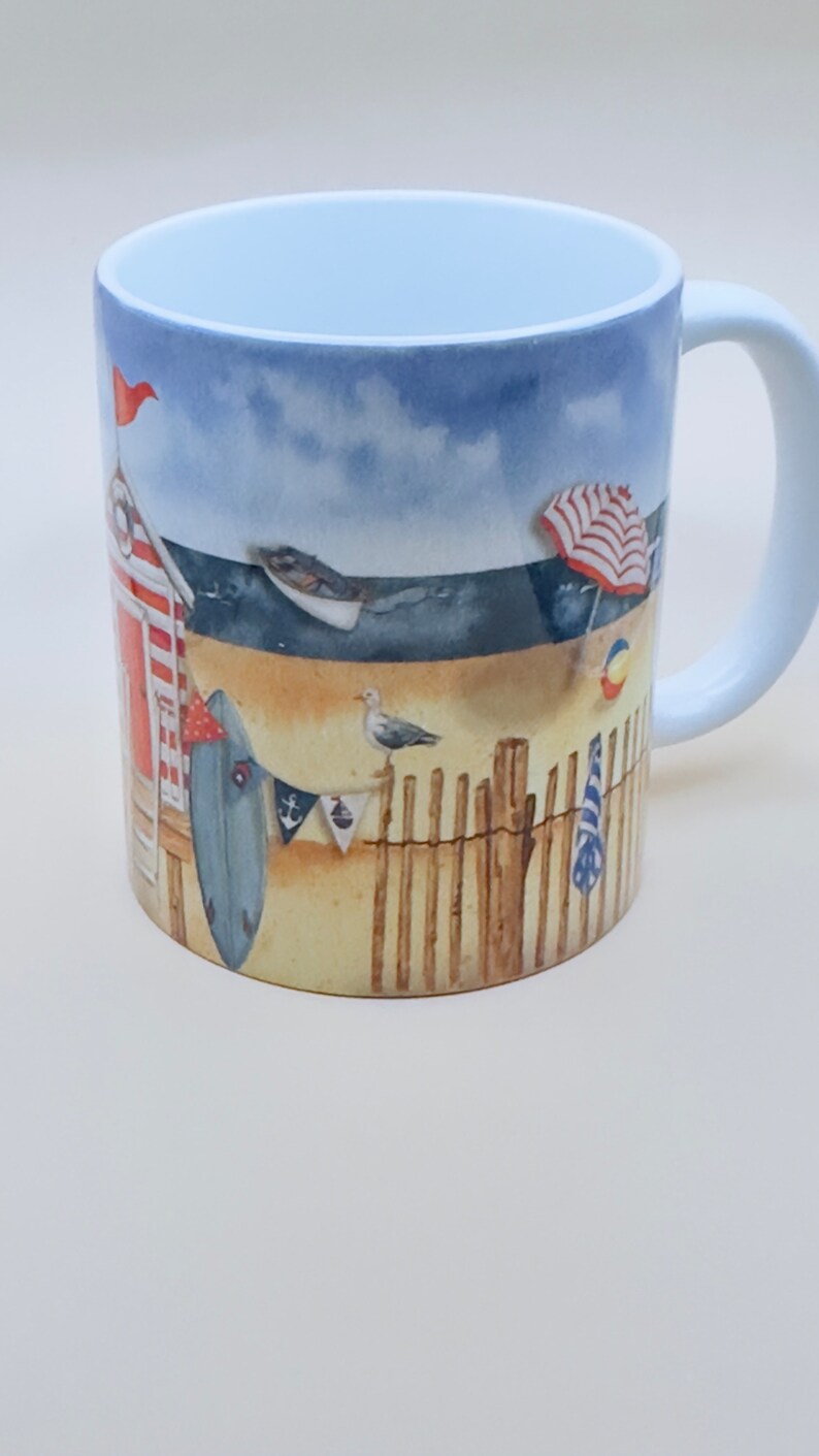 May include: A white ceramic mug with a beach scene design. The mug features a red and white striped beach hut, a blue surfboard, a red and white striped umbrella, a blue and white striped towel, and a seagull. The background is a blue sky with white clouds.
