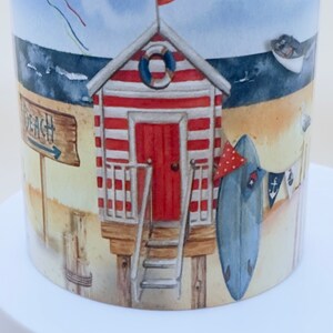 May include: A white ceramic mug with a beach scene design. The mug features a red and white striped beach hut with a red flag, a surfboard, and a sign that says "Beach".