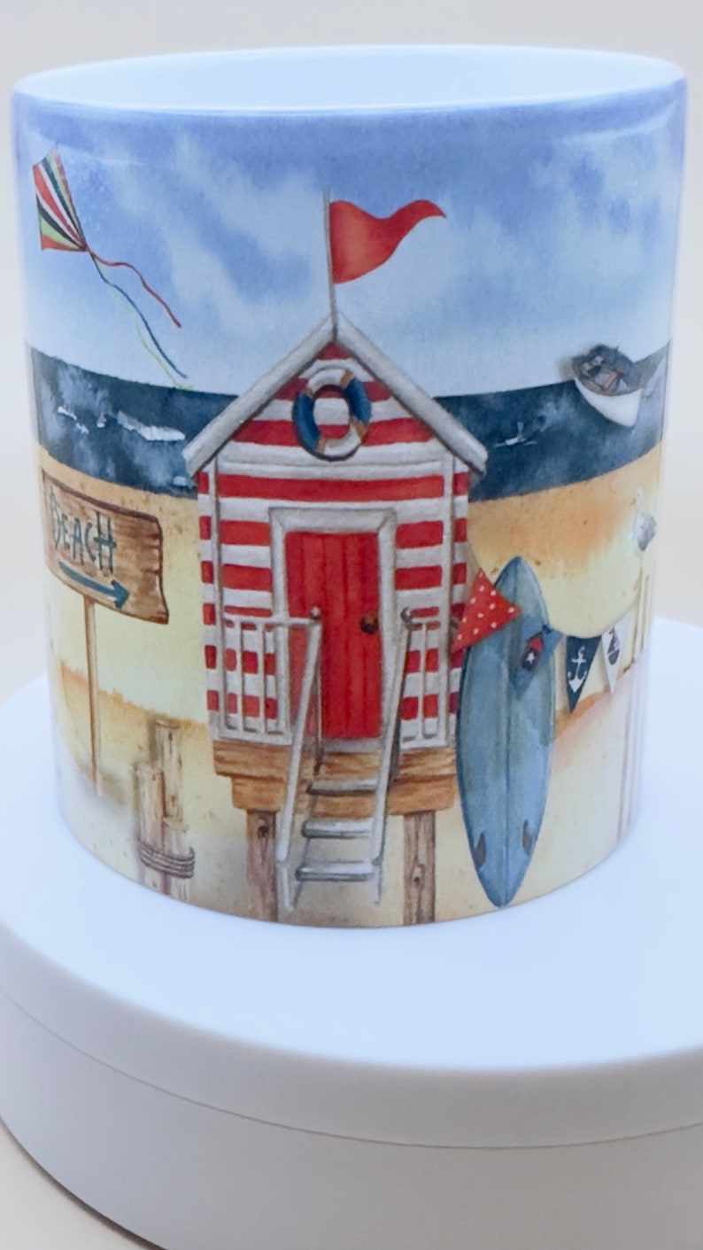 May include: A white ceramic mug with a beach scene design. The mug features a red and white striped beach hut with a red flag, a surfboard, and a sign that says "Beach".