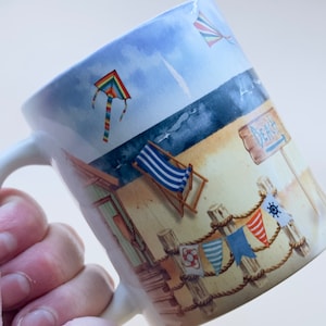 May include: White ceramic mug with a beach scene design. The mug features a beach chair, a beach hut, a kite, and a sign that says "Beach".