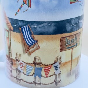 May include: A white ceramic mug with a beach scene design. The mug features a beach hut, a deckchair, a sign that says "Beach", and two kites in the sky. The mug is decorated with a rope and colourful flags.