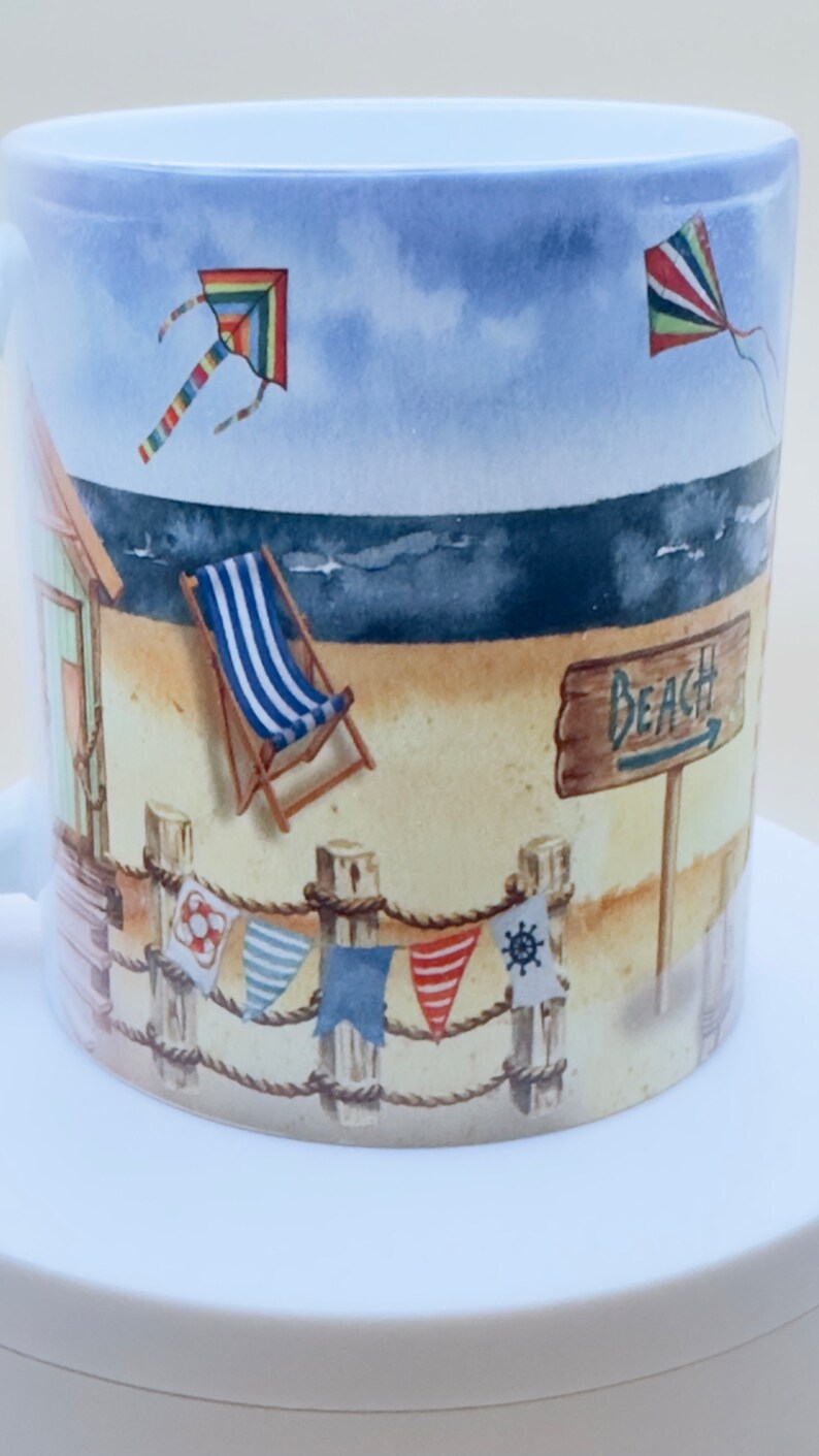 May include: A white ceramic mug with a beach scene design. The mug features a beach hut, a deckchair, a sign that says "Beach", and two kites in the sky. The mug is decorated with a rope and colourful flags.