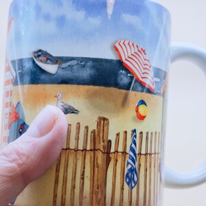 May include: White ceramic mug with a beach scene design. The mug features a blue sky, ocean, sand, a red and white striped beach umbrella, a wooden fence, a beach towel, a beach ball, and a seagull.