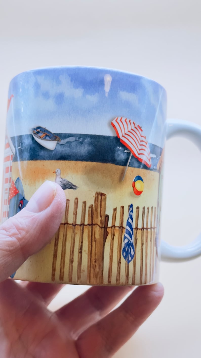 May include: White ceramic mug with a beach scene design. The mug features a blue sky, ocean, sand, a red and white striped beach umbrella, a wooden fence, a beach towel, a beach ball, and a seagull.
