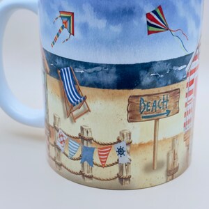 May include: White ceramic mug with a beach scene design. The design features a beach with a deckchair, a sign that says "Beach", and two kites in the sky. There are also colourful flags hanging from a rope.