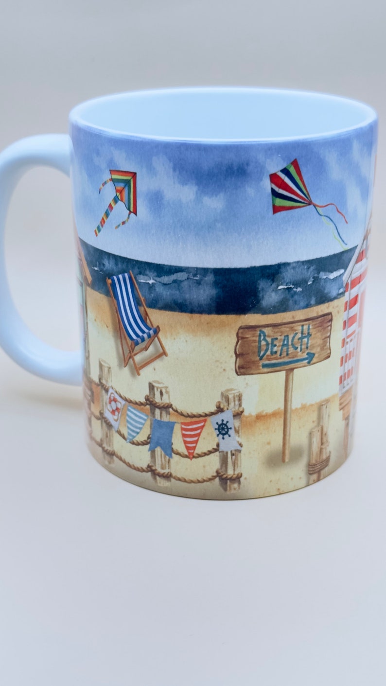 May include: White ceramic mug with a beach scene design. The design features a beach with a deckchair, a sign that says "Beach", and two kites in the sky. There are also colourful flags hanging from a rope.