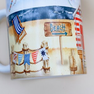 May include: A white ceramic mug with a beach scene design. The mug features a red and white striped beach hut, a wooden sign that says "Beach", a blue and white striped deckchair, and two kites in the sky.