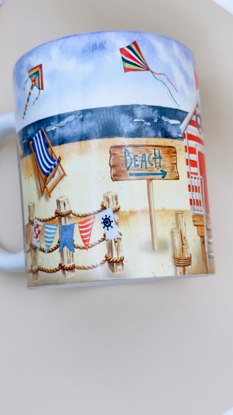 May include: A white ceramic mug with a beach scene design. The mug features a red and white striped beach hut, a wooden sign that says "Beach", a blue and white striped deckchair, and two kites in the sky.