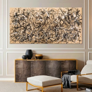 May include: A black and white abstract painting hangs on a wall in a modern living room. The painting is framed and is above a brown cabinet with a white ottoman in front of it. There is a white armchair with a black throw blanket on it to the right of the ottoman.