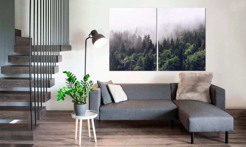 May include: A grey sectional sofa with a fluffy grey throw pillow in a living room with a wooden staircase, a potted plant, and a floor lamp. A large canvas print of a misty forest hangs on the wall above the sofa.