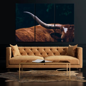 May include: A tri-panel canvas print featuring a close-up of a brown and white longhorn steer with large horns. The steer is set against a dark green background.